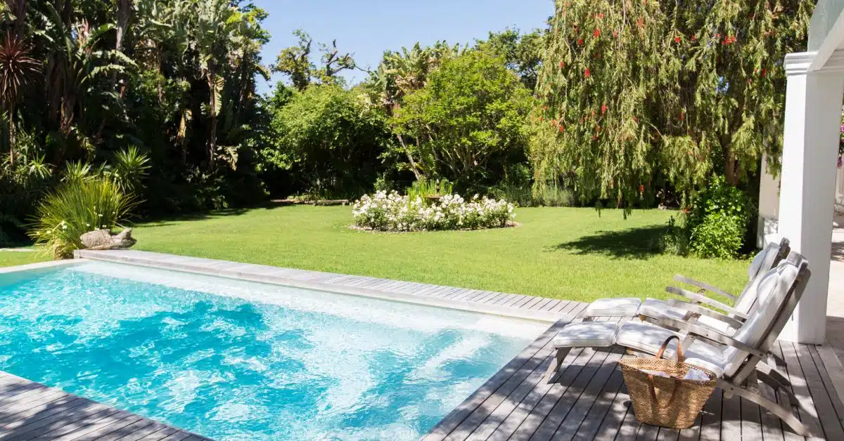 A rectangular pool surrounded by a wooden deck with two lounge chairs on it and grass and plants in the background.