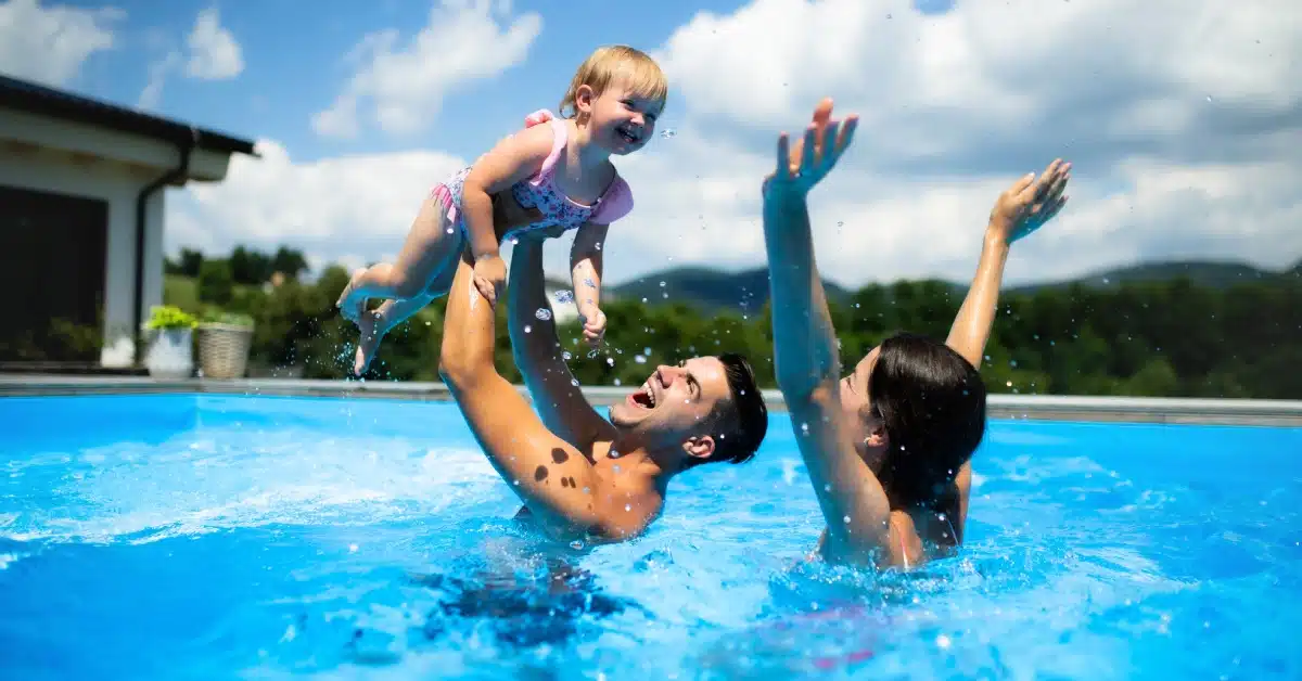 A young couple is holding their baby daughter in the air as they swim in a pool with a house and mountains in the back.