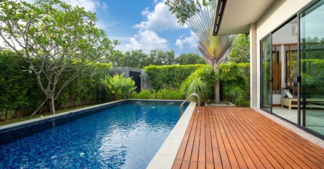 A modern pool next to a wooden deck in the backyard of someone’s home is surrounded by bushes and tropical plants.