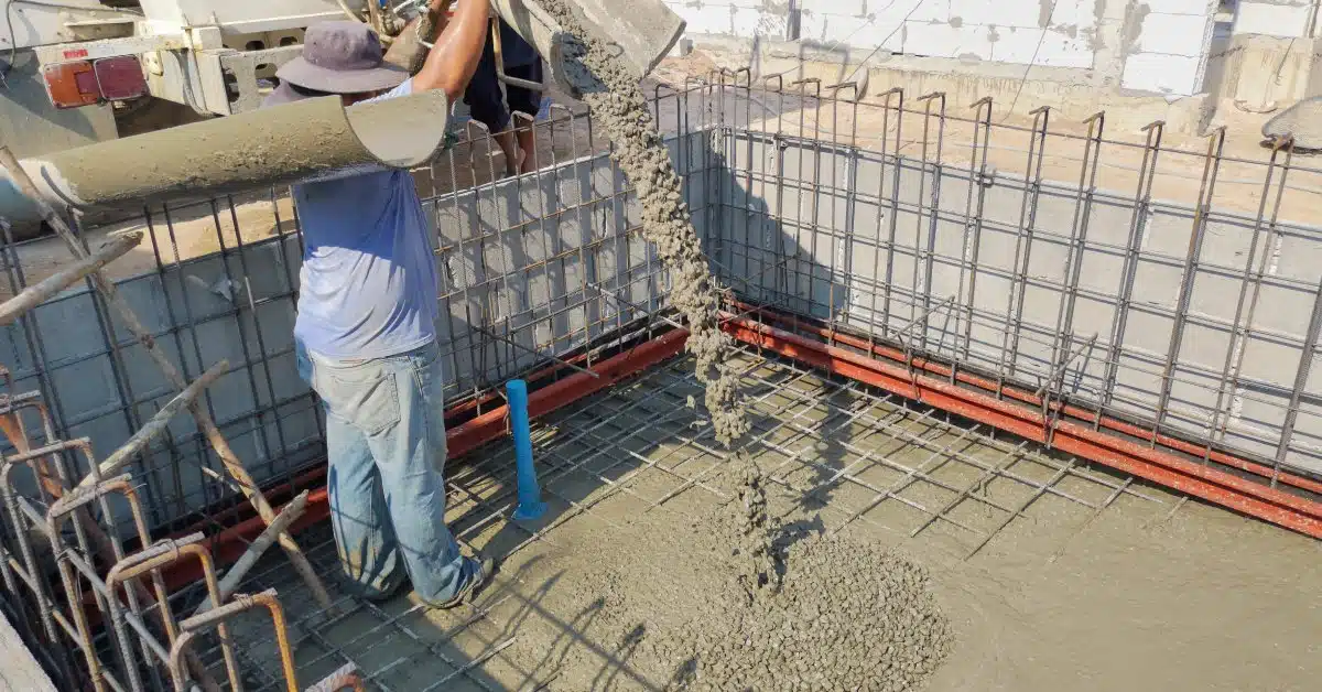 A man in a bucket hat holds a ramp that is pouring liquid concrete into the bottom of a pool under construction.