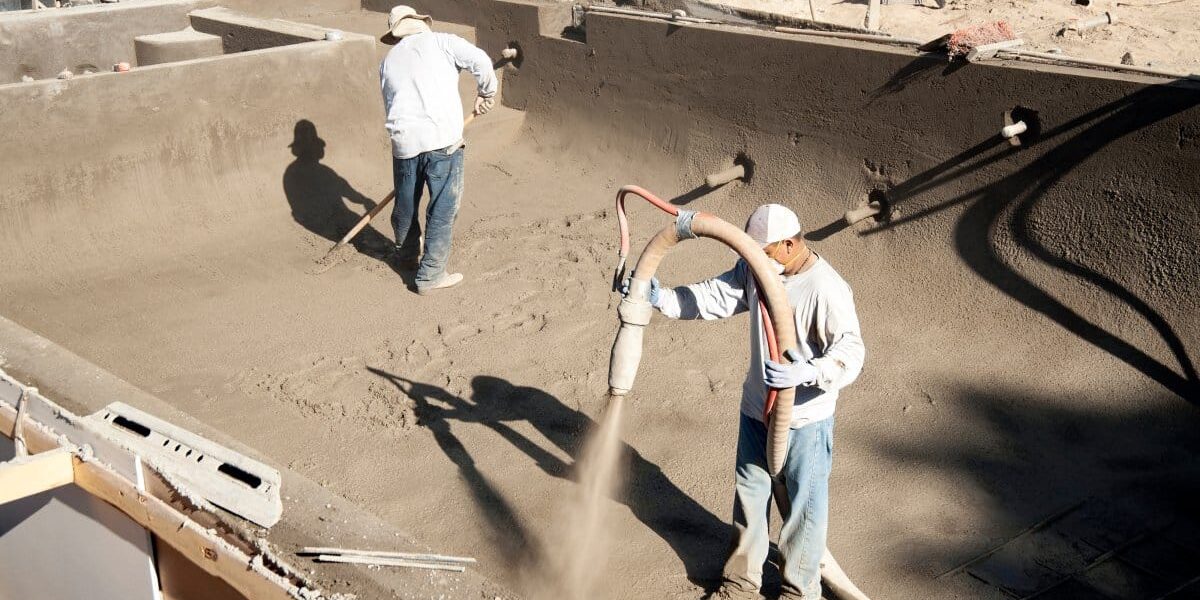 Two pool construction professionals work on putting the foundation of a new pool down using pipes and rakes.