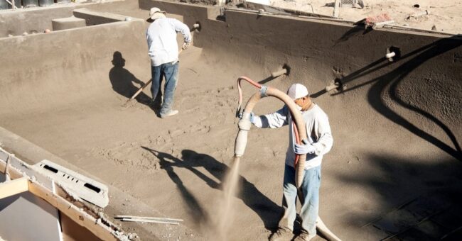 Two pool construction professionals work on putting the foundation of a new pool down using pipes and rakes.