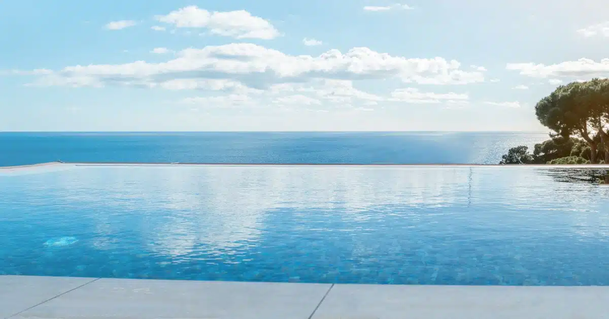 An infinity pool in front of a tiled deck. There are some trees and the ocean beyond the edge of the pool.