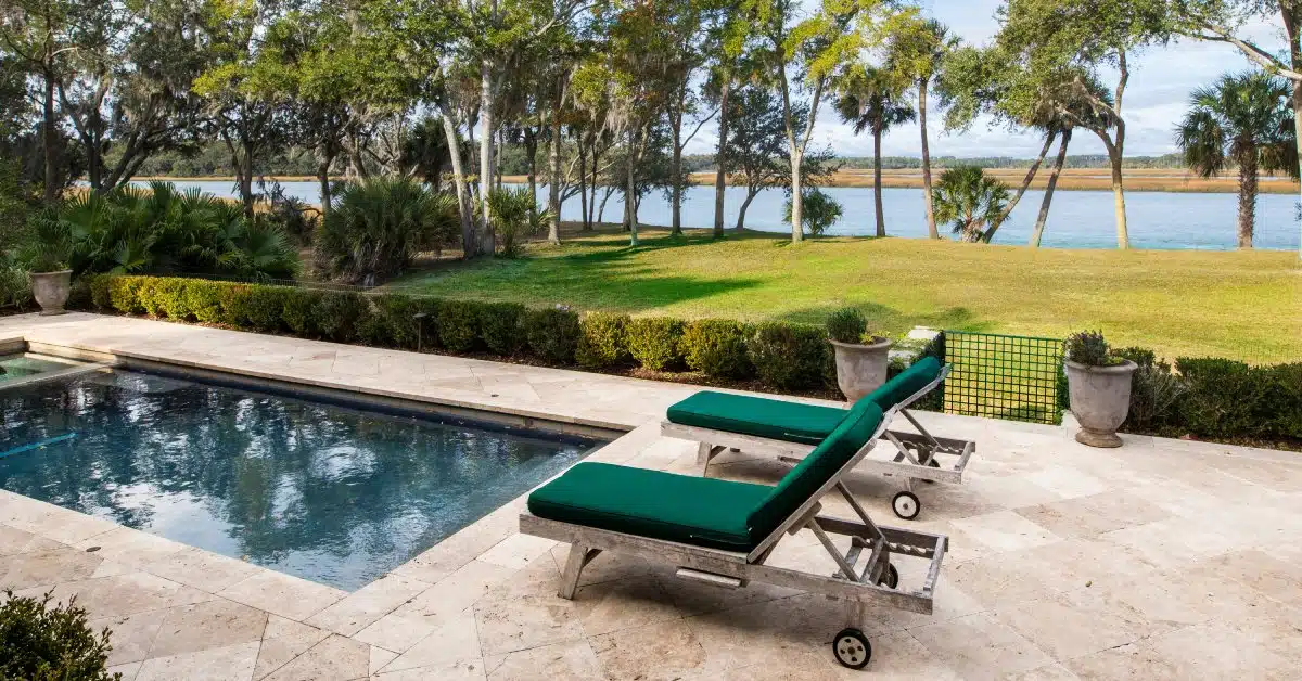 A small pool surrounded by a stone patio. Two green chairs are next to the pool and a green yard stretches beyond it.