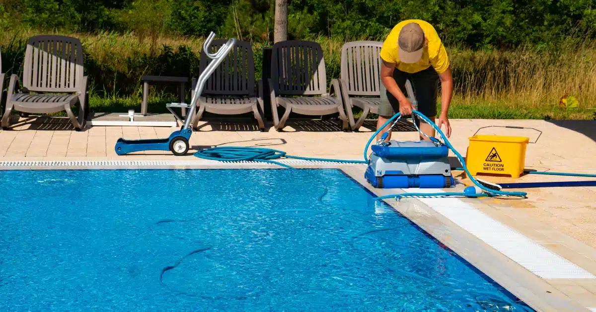 A man in a yellow shirt and a tan hat uses an automatic cleaning robot to clean a large swimming pool.