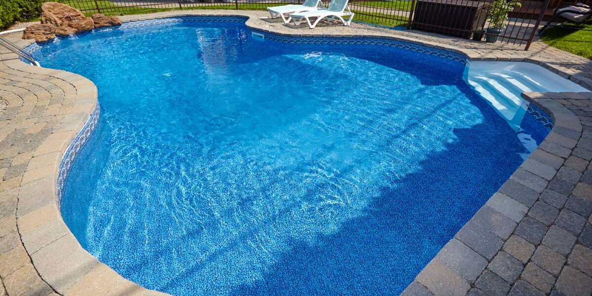 Closeup of a large, kidney-shaped pool surrounded by stone pavers and two lounge chairs in a backyard. Closeup of a large, kidney-shaped pool surrounded by stone pavers and two lounge chairs in a backyard.