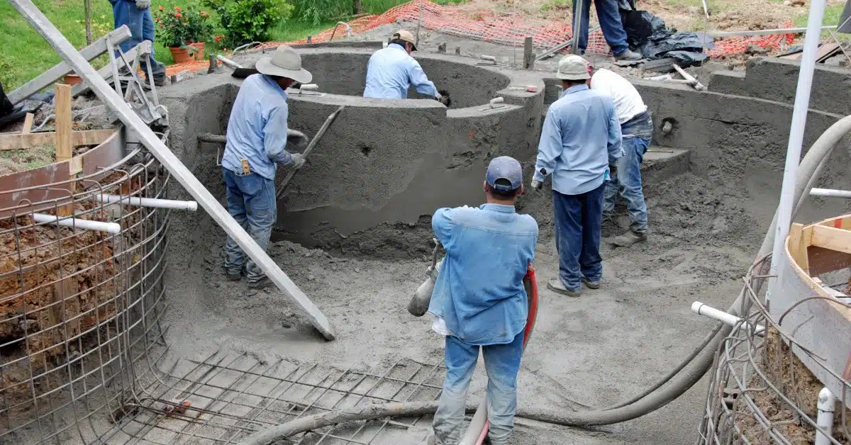 A group of several men in jeans is working on building a pool with a jacuzzi in the backyard of someone’s home.