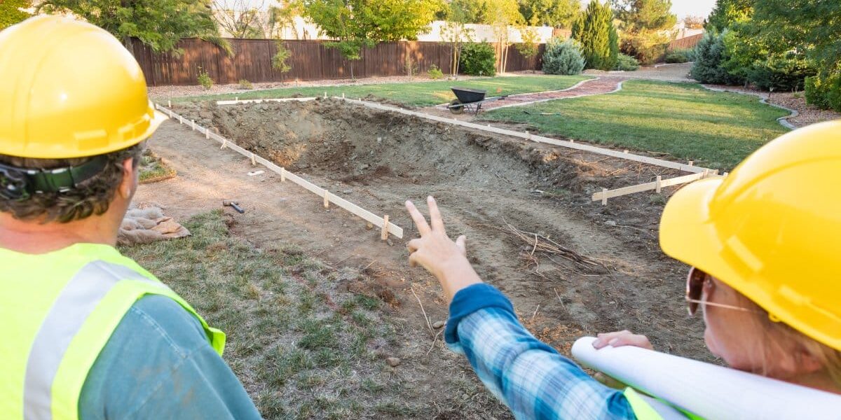 GulfCoastPool-358980-swimming-pool-construction-blogbanner-1 Close-up of the backs of two people wearing hard hats and pointing at the construction site of a swimming pool.