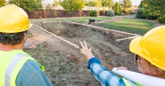 Close-up of the backs of two people wearing hard hats and pointing at the construction site of a swimming pool.