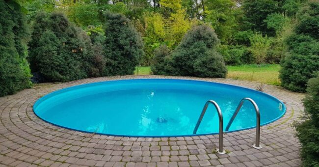 A small round plunge pool with metal stairs surrounded by a stone patio and various bushes and trees.