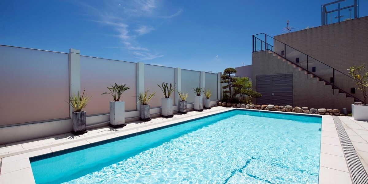 Closeup of a large, rectangular pool surrounded by concrete, a tall fence, potted plants, and a set of stairs.