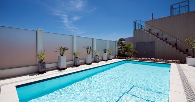 Closeup of a large, rectangular pool surrounded by concrete, a tall fence, potted plants, and a set of stairs.