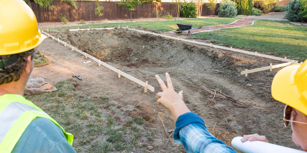 GulfCoastPool-365130-two-pool-contractors-blogbanner-1 Rear view of a man and woman in yellow hard hats stand talking while pointing at the construction of a backyard pool.