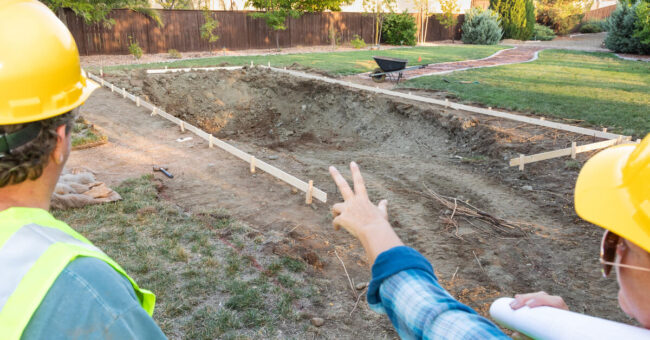 Rear view of a man and woman in yellow hard hats stand talking while pointing at the construction of a backyard pool.