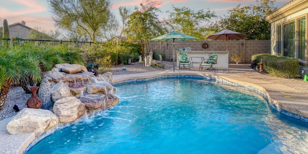 A custom pool with a waterfall feature surrounded by landscaping in a residential backyard with an iron fence.