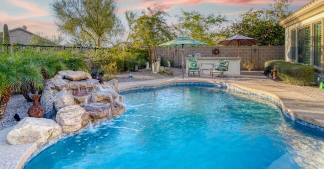 A custom pool with a waterfall feature surrounded by landscaping in a residential backyard with an iron fence.