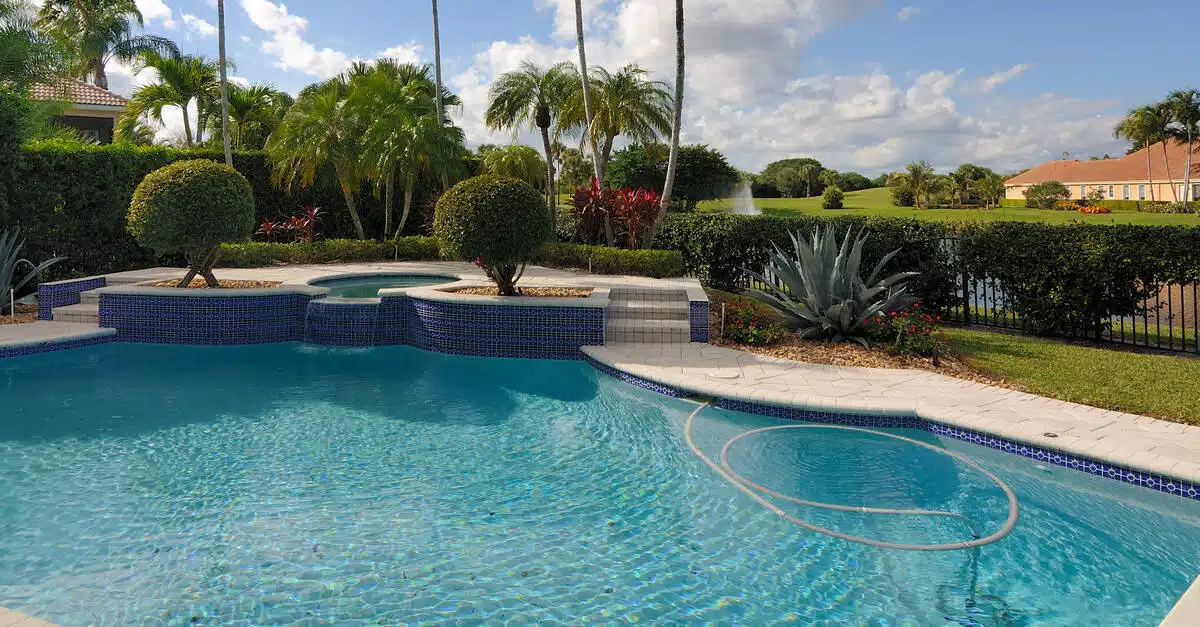 A custom pool with a jacuzzi in a backyard with several palm trees, manicured bushes and tropical plants.