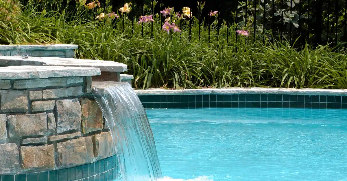A tiled pool with a cascading stone waterfall surrounded by tall grasses and purple and yellow flowers.