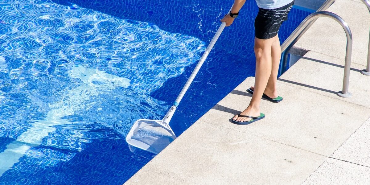 An individual in a T-shirt, shorts, and flip flops using a net with a long handle to clean a swimming pool.