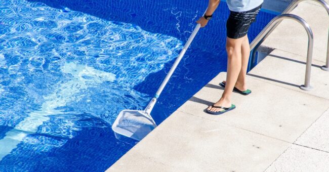 An individual in a T-shirt, shorts, and flip flops using a net with a long handle to clean a swimming pool.