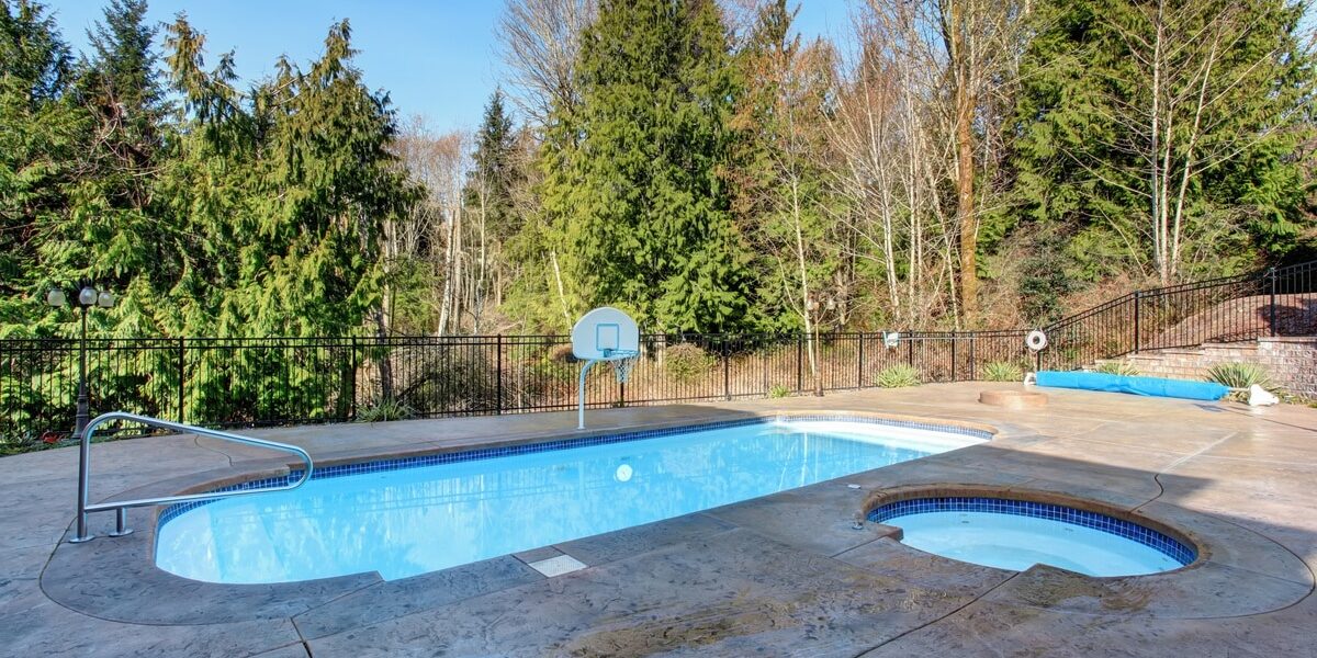 An oval-shaped pool with a basketball hoop and a circular jacuzzi surrounded by a concrete deck and mature trees. An oval-shaped pool with a basketball hoop and a circular jacuzzi surrounded by a concrete deck and mature trees.