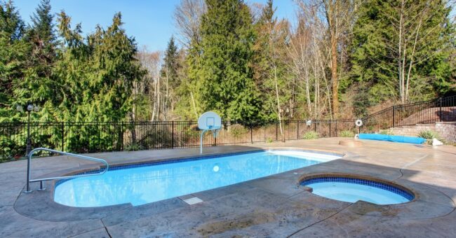 An oval-shaped pool with a basketball hoop and a circular jacuzzi surrounded by a concrete deck and mature trees.