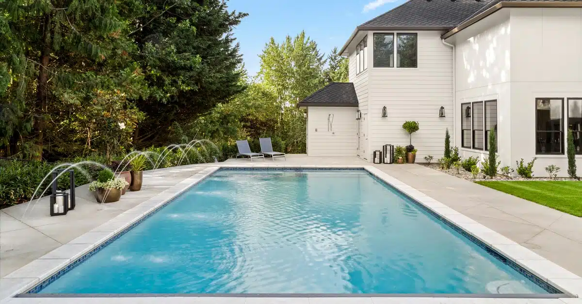 A rectangular pool with water jets forming arches in the backyard of a white multistory house on a sunny day.
