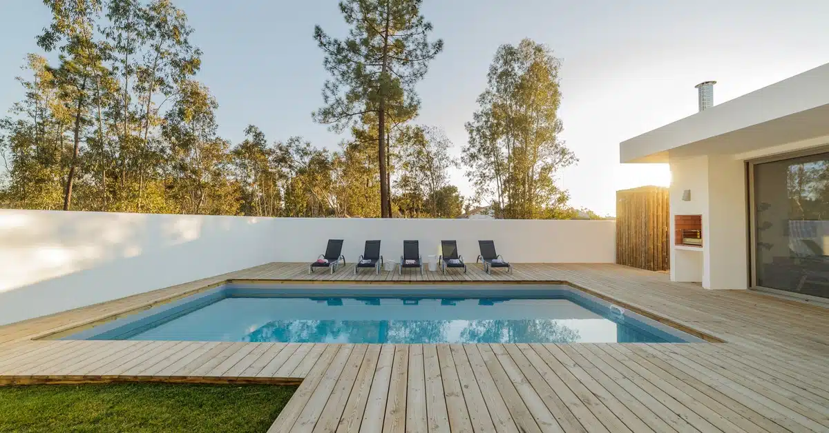 A modern house with a square pool in the backyard surrounded by a wood deck and several lounge chairs.