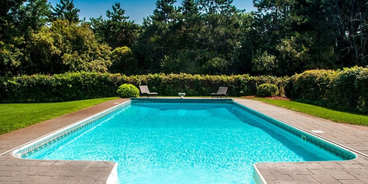 A rectangular outdoor pool with two lounge chairs surrounded by a stone deck, green hedges, and mature trees.