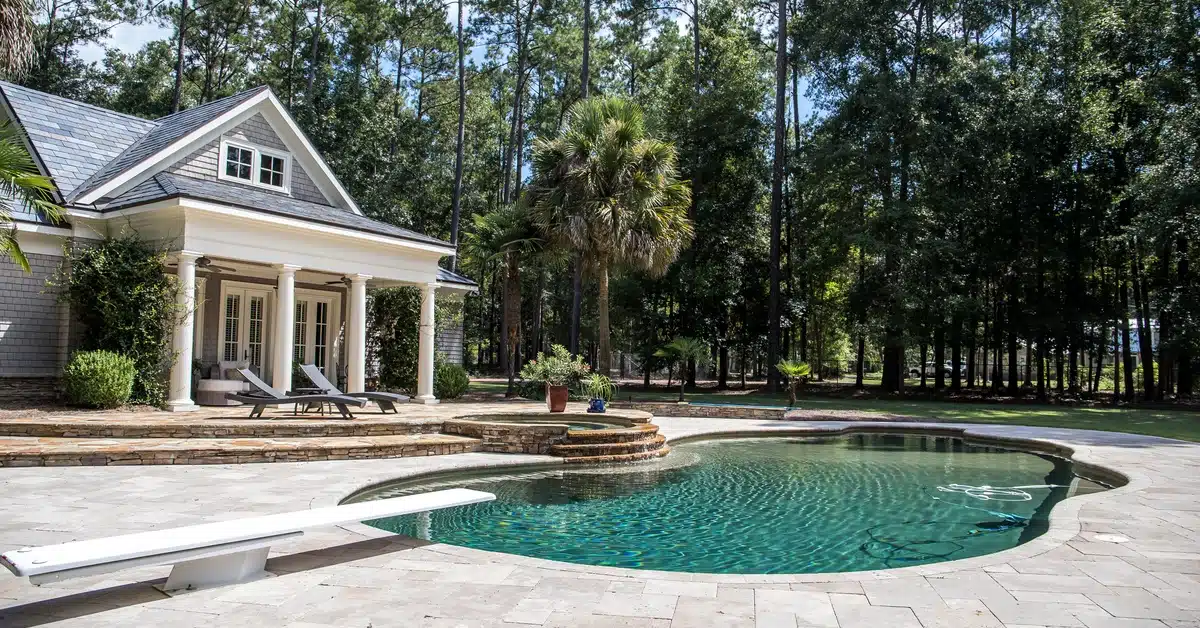 A backyard pool and hot tub with a tiled deck and diving board surrounded by mature trees on a sunny day.