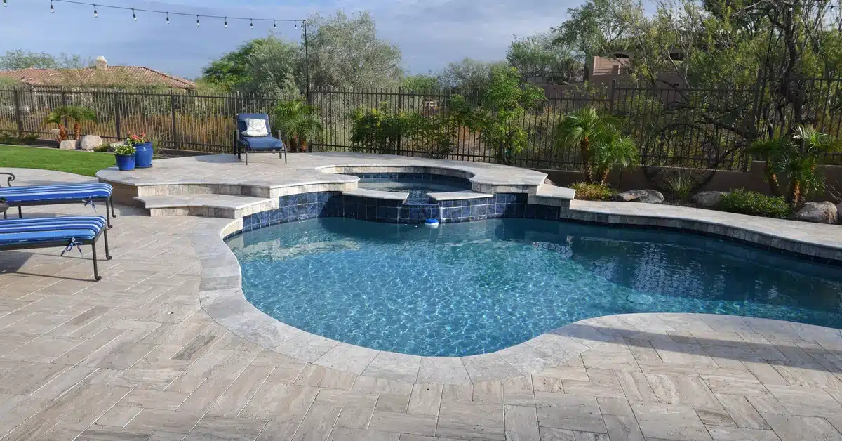 A backyard pool and hot tub with a tiled deck surrounded by plants and sun loungers with blue cushions.