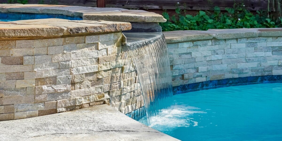 The Advantages of Having a Custom-Built Pool in Your House A pool with custom white brick walls and a cascading water feature connecting a hot tub to the main pool.