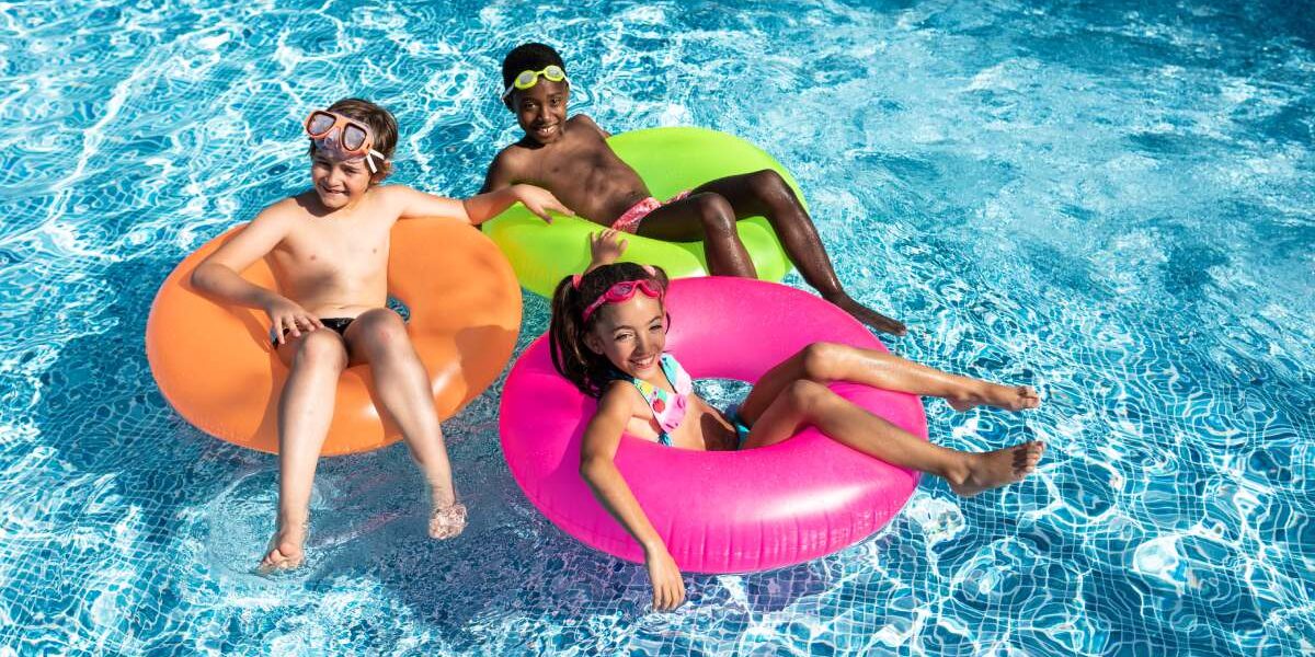 Two smiling boys and a girl wearing bathing suits and goggles while floating in colorful tubes in a pool.