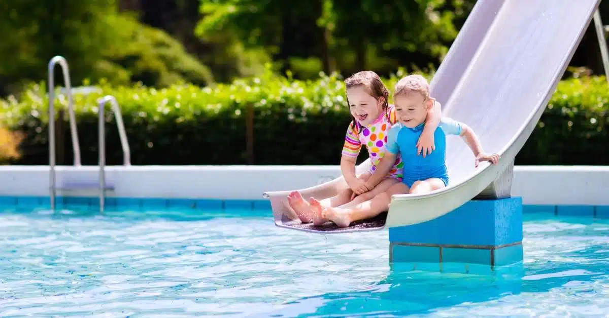 Two smiling young children in bathing suits holding each other as they enjoy an outdoor water slide.