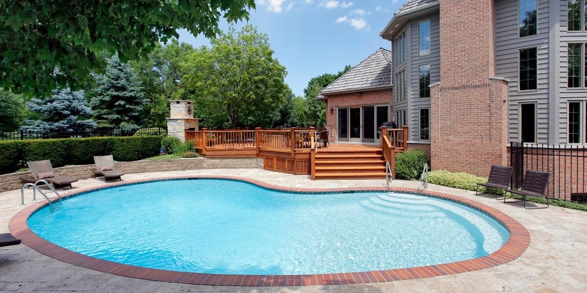 The backyard of a gray multistory house shows a circular swimming pool, a wooden deck, and an outdoor fireplace.