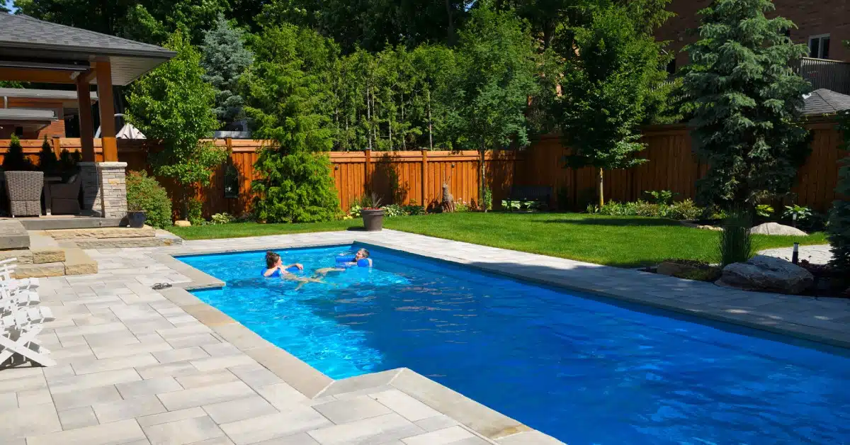 Two people using blue floats in a rectangular swimming pool surrounded by grass, a wooden fence, and trees.