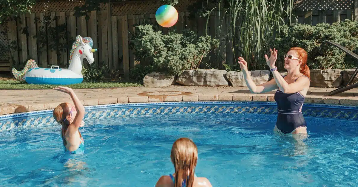 A woman and two young girls standing in a swimming pool while playing with a multicolored beach ball.
