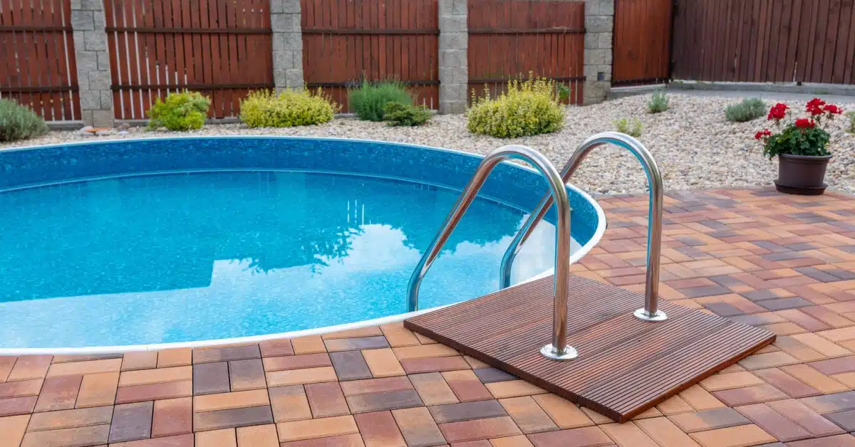 A circular swimming pool with a ladder surrounded by a brick patio, rocks, low-profile plants, and a wood fence.