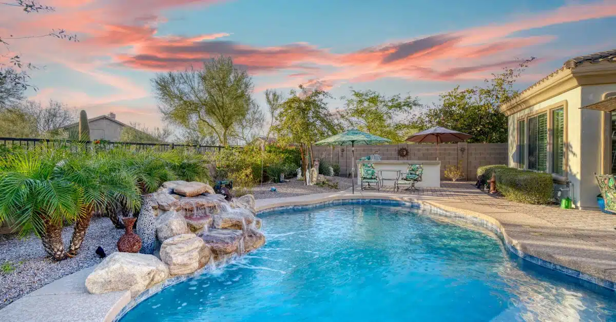 A backyard with a freeform pool with a waterfall surrounded by a patio with two chairs and umbrellas.