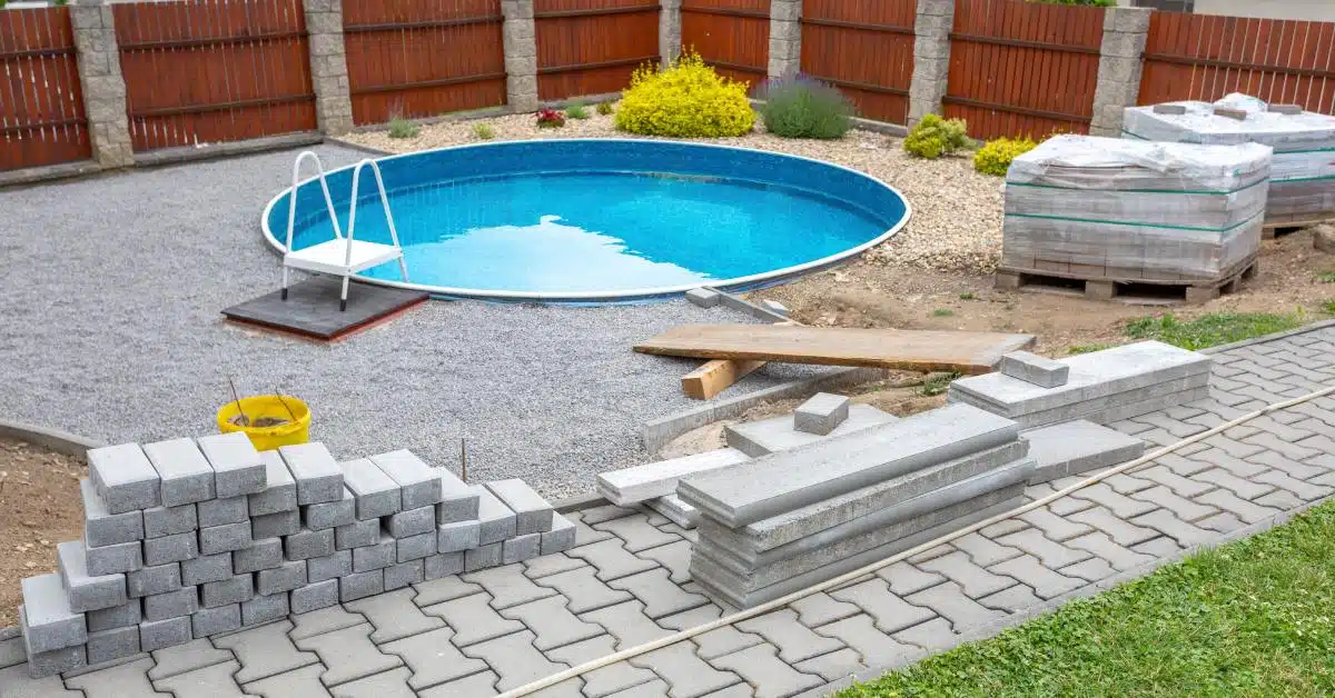 A circular swimming pool that is under construction and surrounded by a fence, landscaping, and a brick walkway.