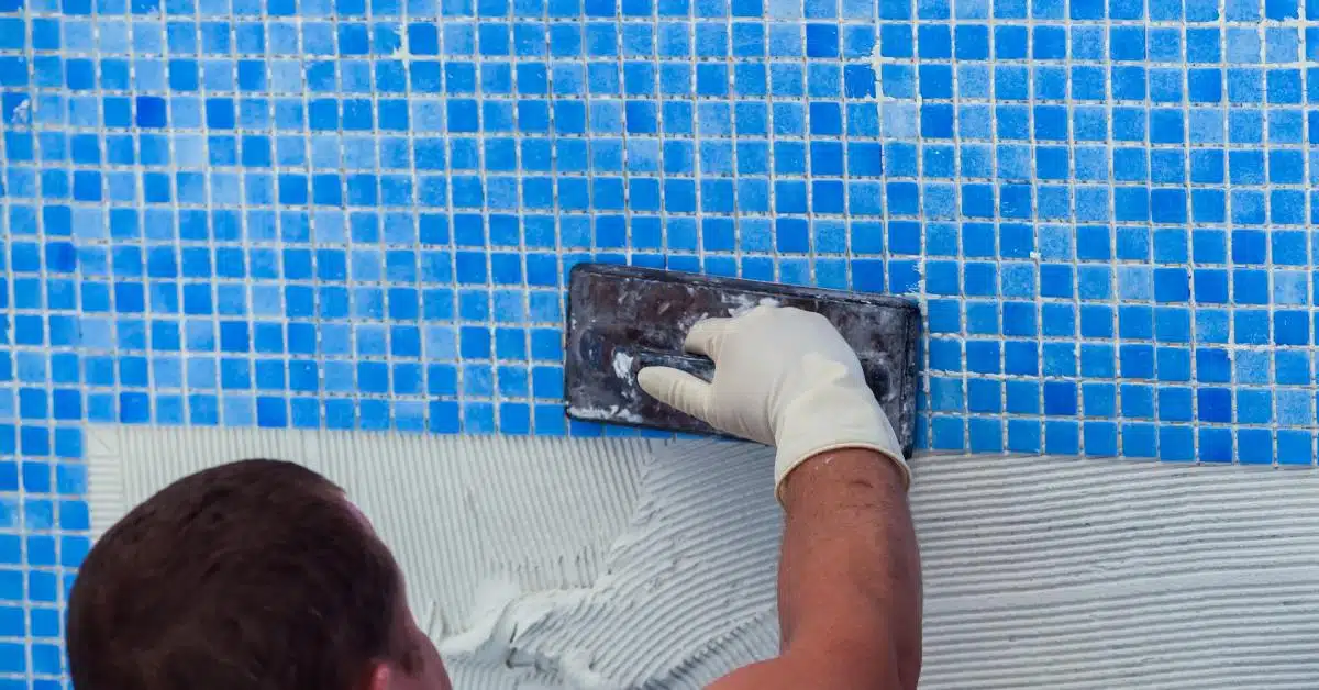 A man in a white T-shirt and gloves using a rectangular trowel and grout to install blue tiles in a pool.