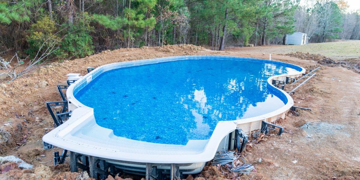 A kidney-shaped swimming pool under construction and filled with water, surrounded by brown dirt and mature trees.
