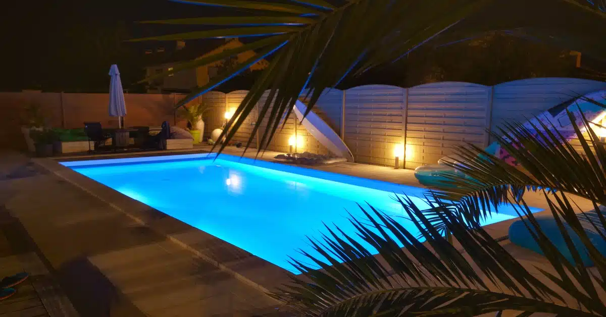 A rectangular pool lit at night and surrounded by a light-colored fence, an outdoor dining area, and potted plants.