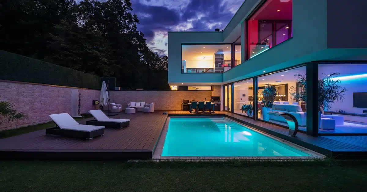 A modern backyard at night, featuring a lit rectangular pool, patio furniture, and an outdoor dining area.