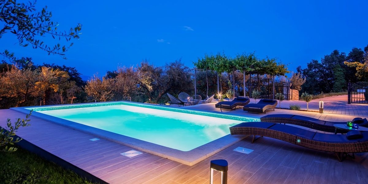 A lit rectangular pool surrounded by composite decking, lounge chairs, grass, and landscaping. The sky above is dark.