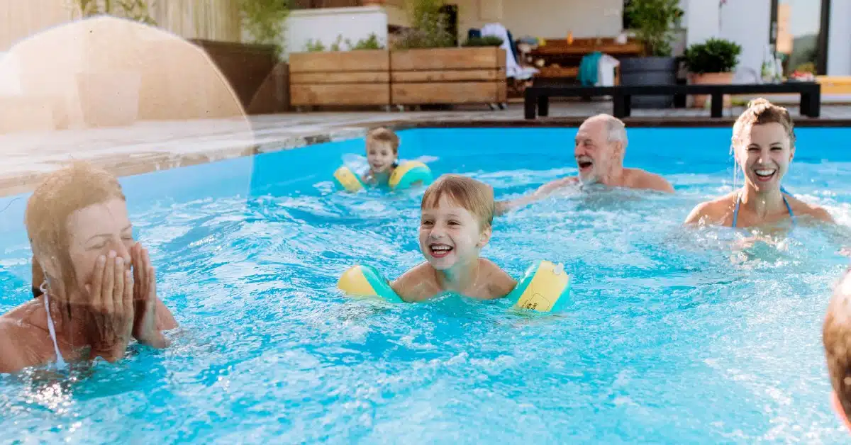 A multi-generational family smiles while swimming in a backyard pool. The children wear yellow and blue arm floats.