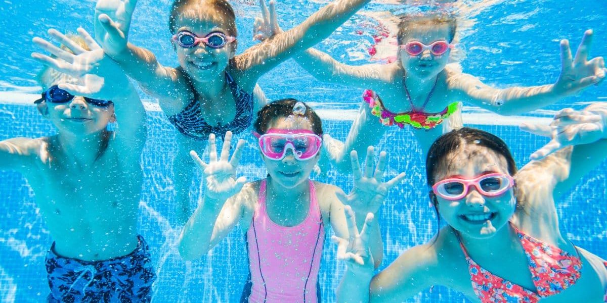 Four girls and one boy in colorful swimsuits and goggles smile as they swim underwater in a pool on a sunny day.