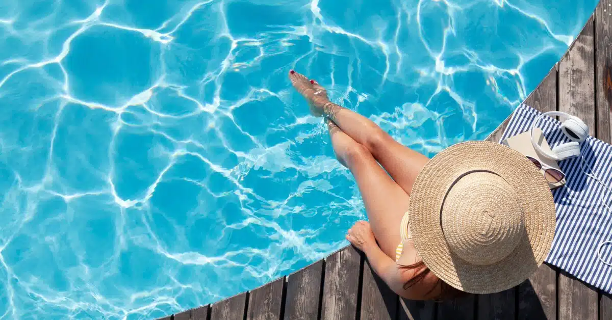 An overhead view of a woman in a swimsuit and large sun hat sitting on a pool deck with her feet in the water.
