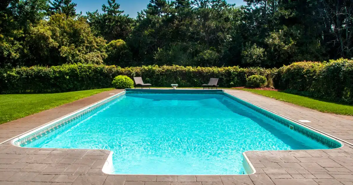 A rectangular swimming pool with a diving board surrounded by two lounge chairs and mature landscaping.
