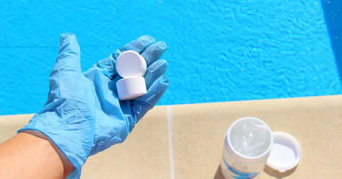 An individual in blue gloves stands next to a pool and holds two white, round chlorine tablets on a sunny day.